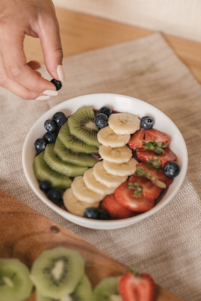 white bowl of fruit with hand placing blueberries on top of kiwi, strawberries, and banana.